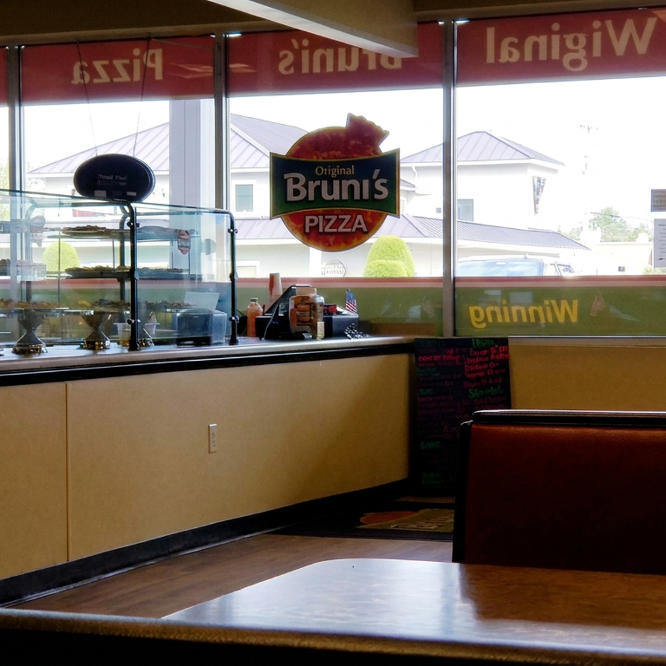 Interior of Bruni’s Pizza restaurant with a display case, condiment bottles, dining booths, and bright front windows.